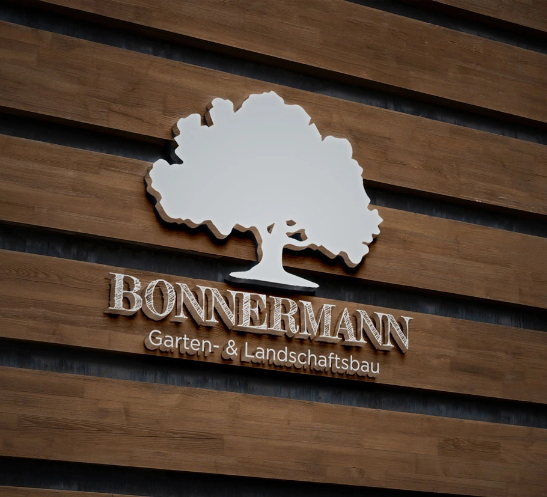 3D-Logo von Bonnermann Garten- & Landschaftsbau an einer Holzfassade montiert, mit stilisiertem Baum und Firmenschriftzug.