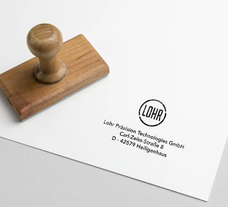 Ein Holzstempel mit rundem Griff neben einem Abdruck auf weißem Papier, der „Lohr Präzision Technologies GmbH, Carl-Zeiss-Straße 8, D-42579 Heiligenhaus“ zeigt. Der Stempelabdruck enthält den Firmennamen „LOHR“ in Fettdruck, umrahmt von einem minimalistischen Design.